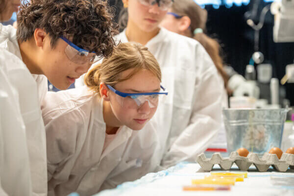 A boy and girl in a lab coat and goggles watch an expiriment.