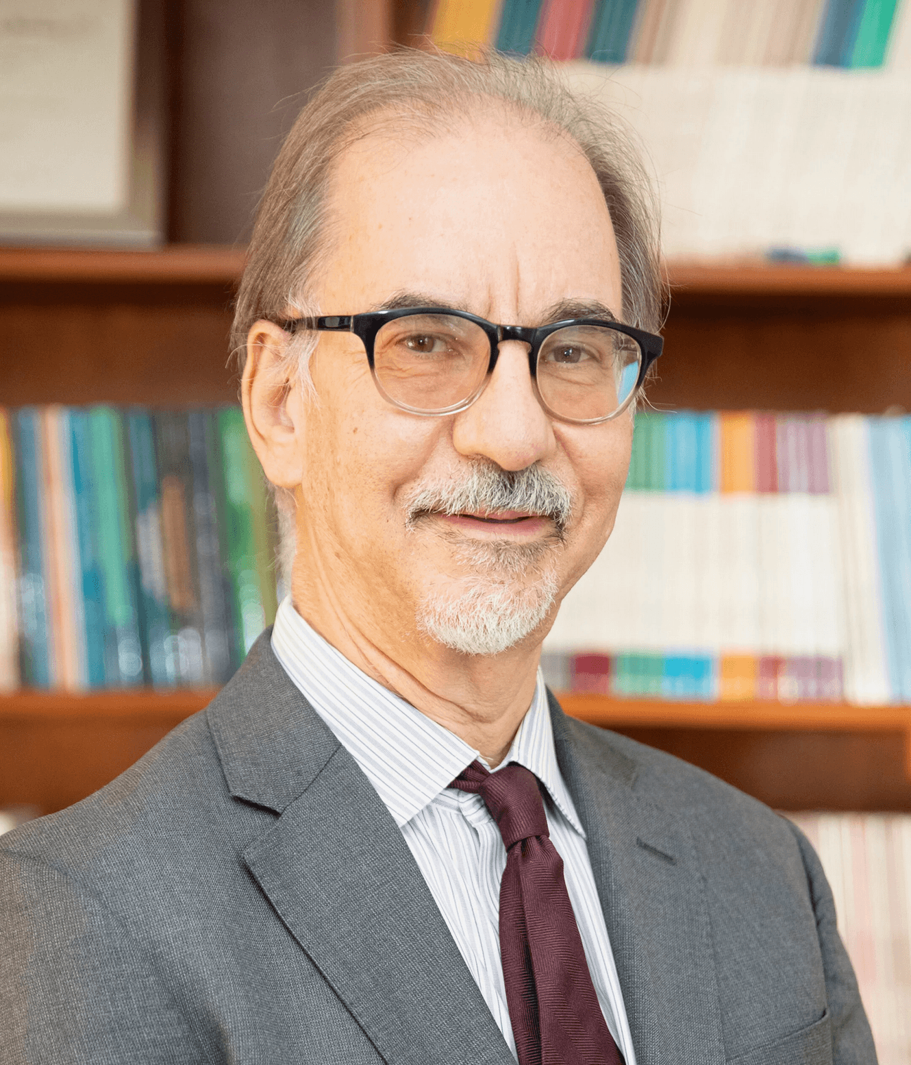 A man with glasses, wearing a grey suit and red tie, stands in front of a bookshelf.