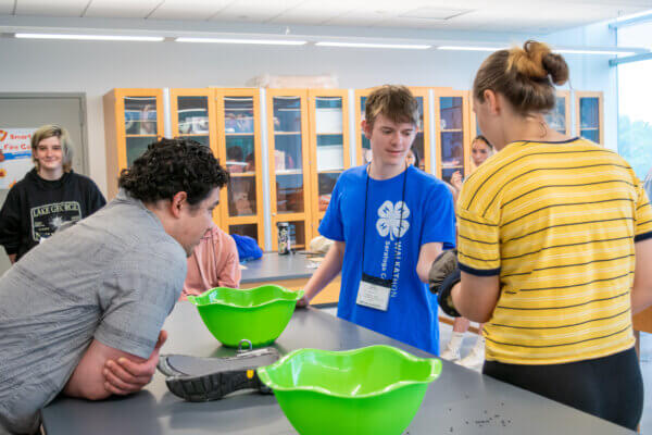 Two teens thumbwrestle while wearing firefighter gloves while a person looks on