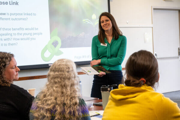 A woman in a green long-sleeved shirt stands at the front of the room listening to three people who have their backs to the camera.