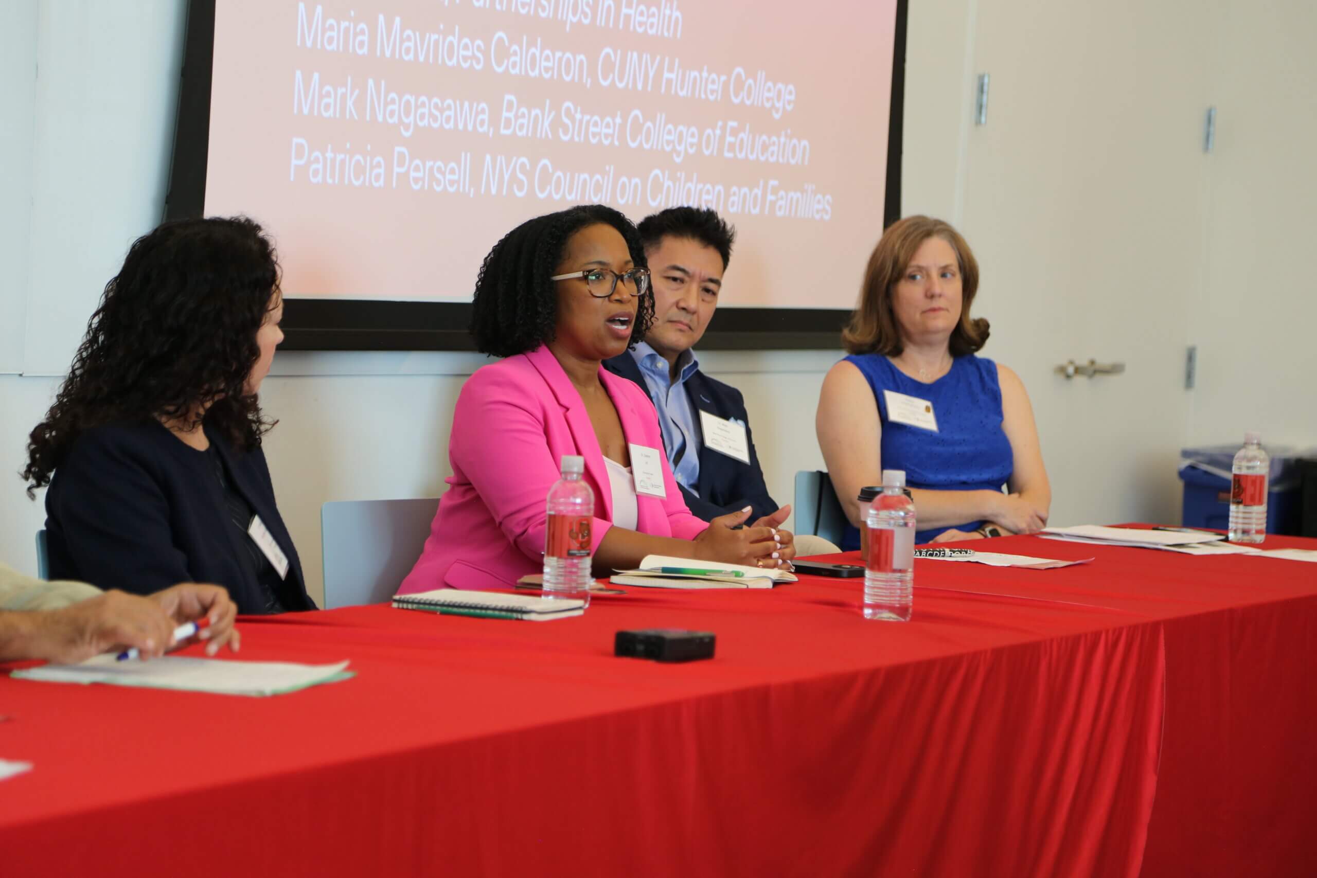 A woman in a pink blazer sits at a table with three other people and gives a lecture.