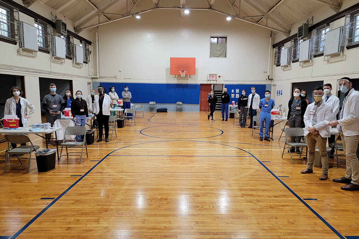 a gym lined with tables where people in medical gear wearing masks stand